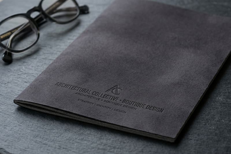 Gemini said Dark grey suede notebook with an embossed architectural logo next to a pair of glasses.