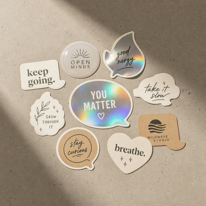 Eight custom sticker samples in matte gloss clear foil holographic and kraft finishes arranged on warm light concrete surface