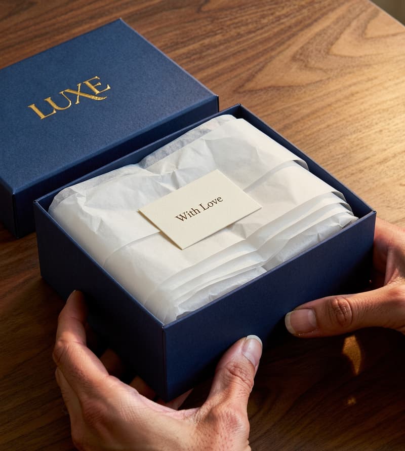 Hands opening a navy branded gift box revealing tissue paper with a gold logo print and a personalised insert card