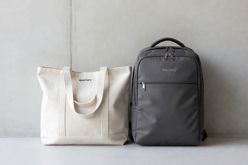 Branded canvas tote bag and structured laptop backpack with small embroidered logo against a concrete wall