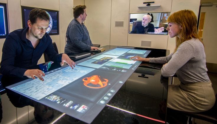 This may contain: three people are sitting at a table with a large screen on it, and one person is standing in front of them