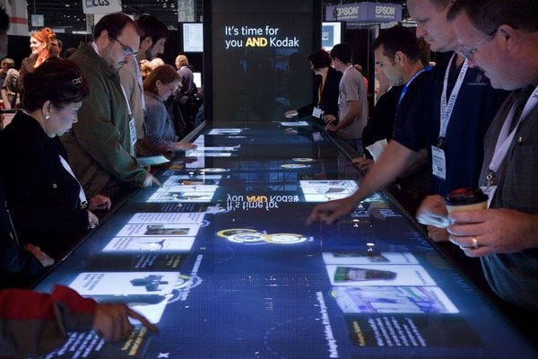 This may contain: a group of people standing around a long table with electronic screens on top of it