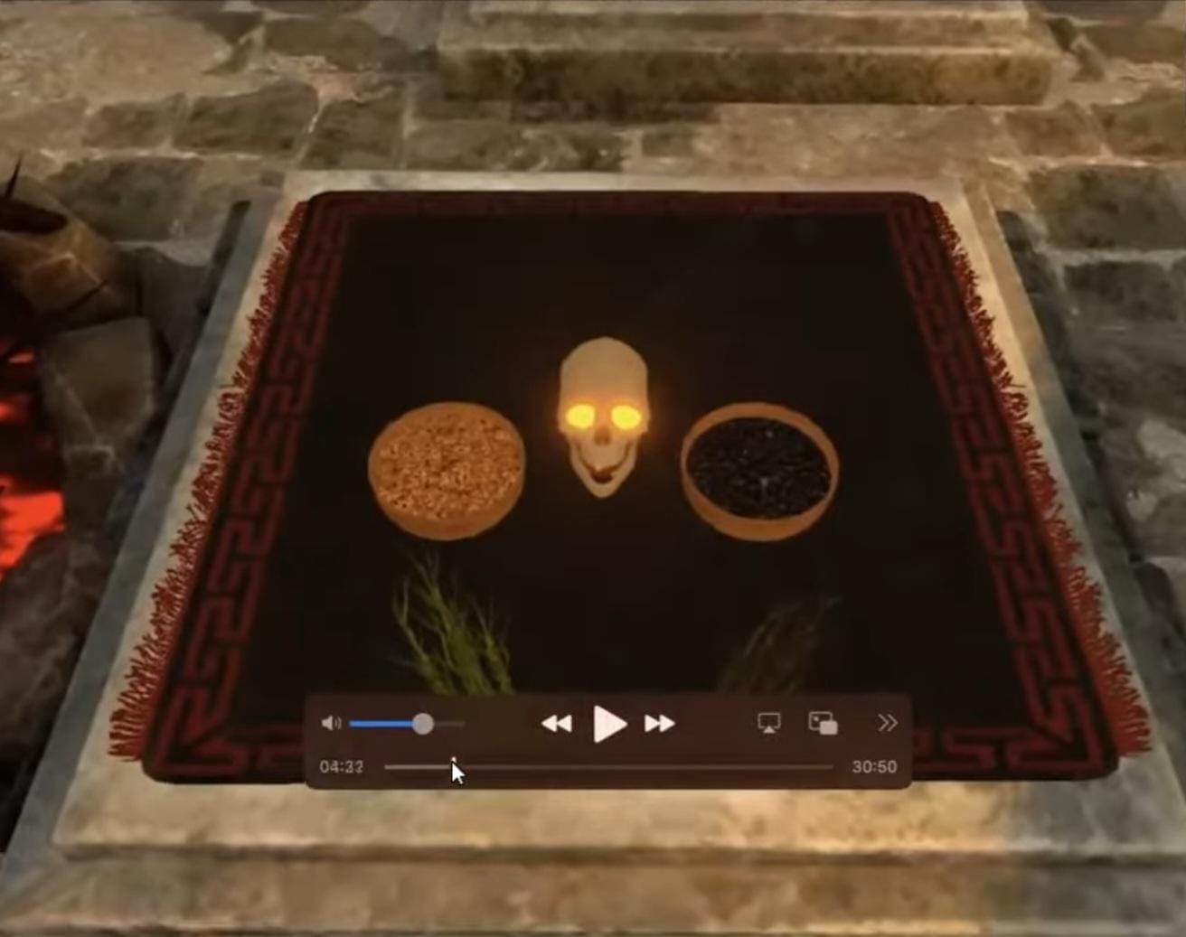 The image is a screenshot of the virtual 3D Kapal Puja that can be performed in the Tantra Sadhana app. It shows a human skull lit with a Diya from within, placed on a red carpet on the floor. Around it are various offerings like grains or lentils in 2 bowls and 2 bunches of herbs or leaves.
