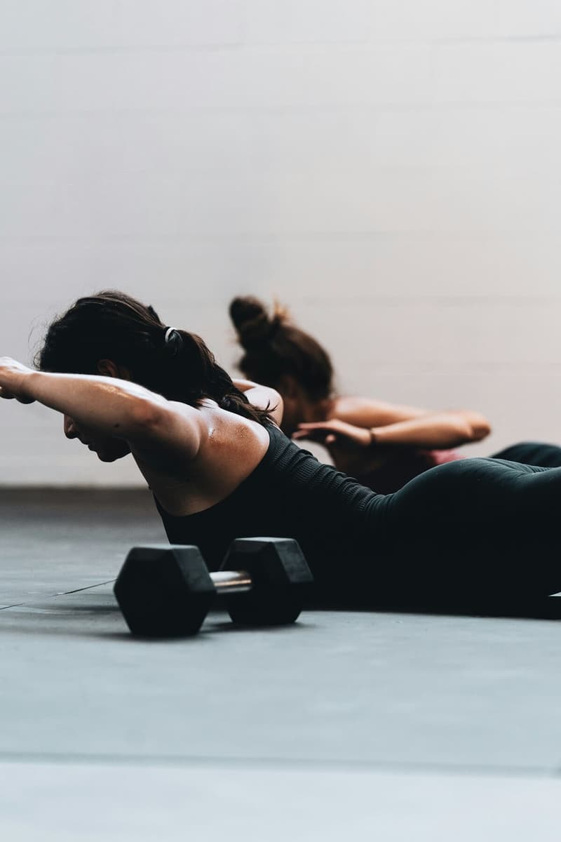Two women performing the superman back extension exercise on a gym floor, with a black hex dumbbell blurred in the foreground.