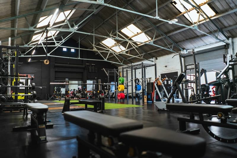 Interior view of a modern functional fitness facility featuring weight training benches, weight plates on a storage rack, a turf strip, and workout rigs under a high-exposed industrial roof.