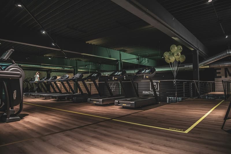 A spacious gym interior featuring a row of modern treadmills lined up on a wooden floor in a cardio zone with industrial-style lighting.