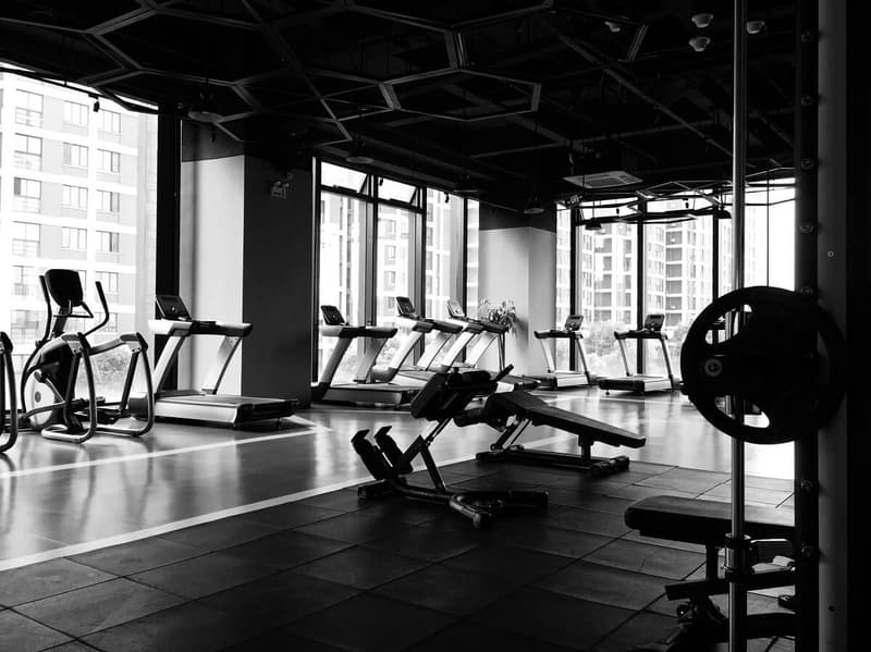 A high-contrast black and white shot of an empty fitness center with cardio equipment, strength training benches, and a honeycomb-patterned ceiling overlooking city buildings.