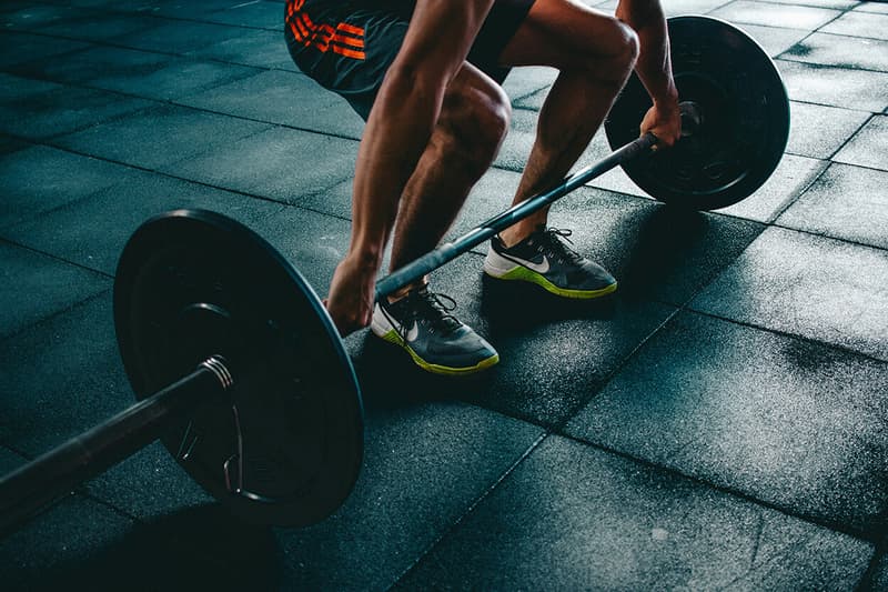 Close-up view of an athlete's legs and hands gripping a barbell in a deadlift starting position inside a gym